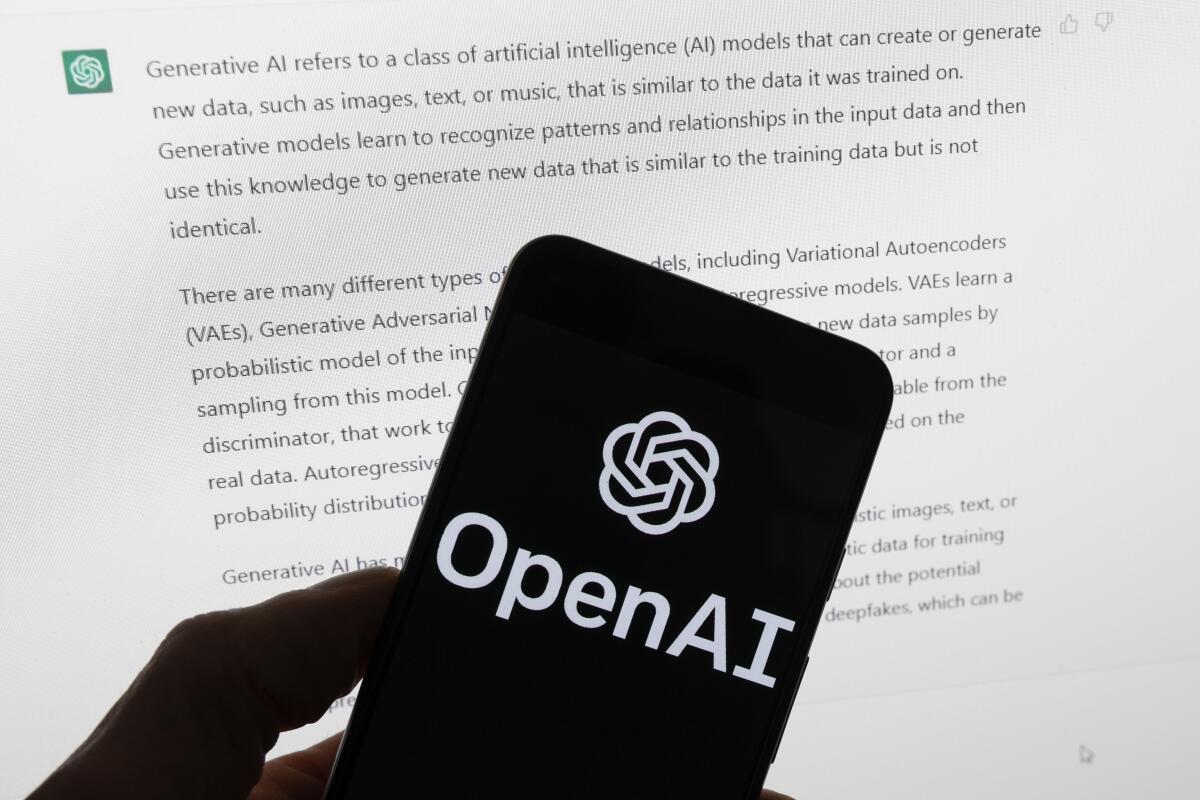 The OpenAI logo is seen on a mobile phone in front of a computer screen displaying output from ChatGPT.