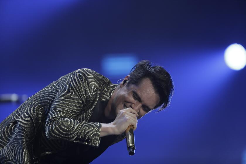 A man with short, dark hair in a patterned suit hunching over while singing into a microphone