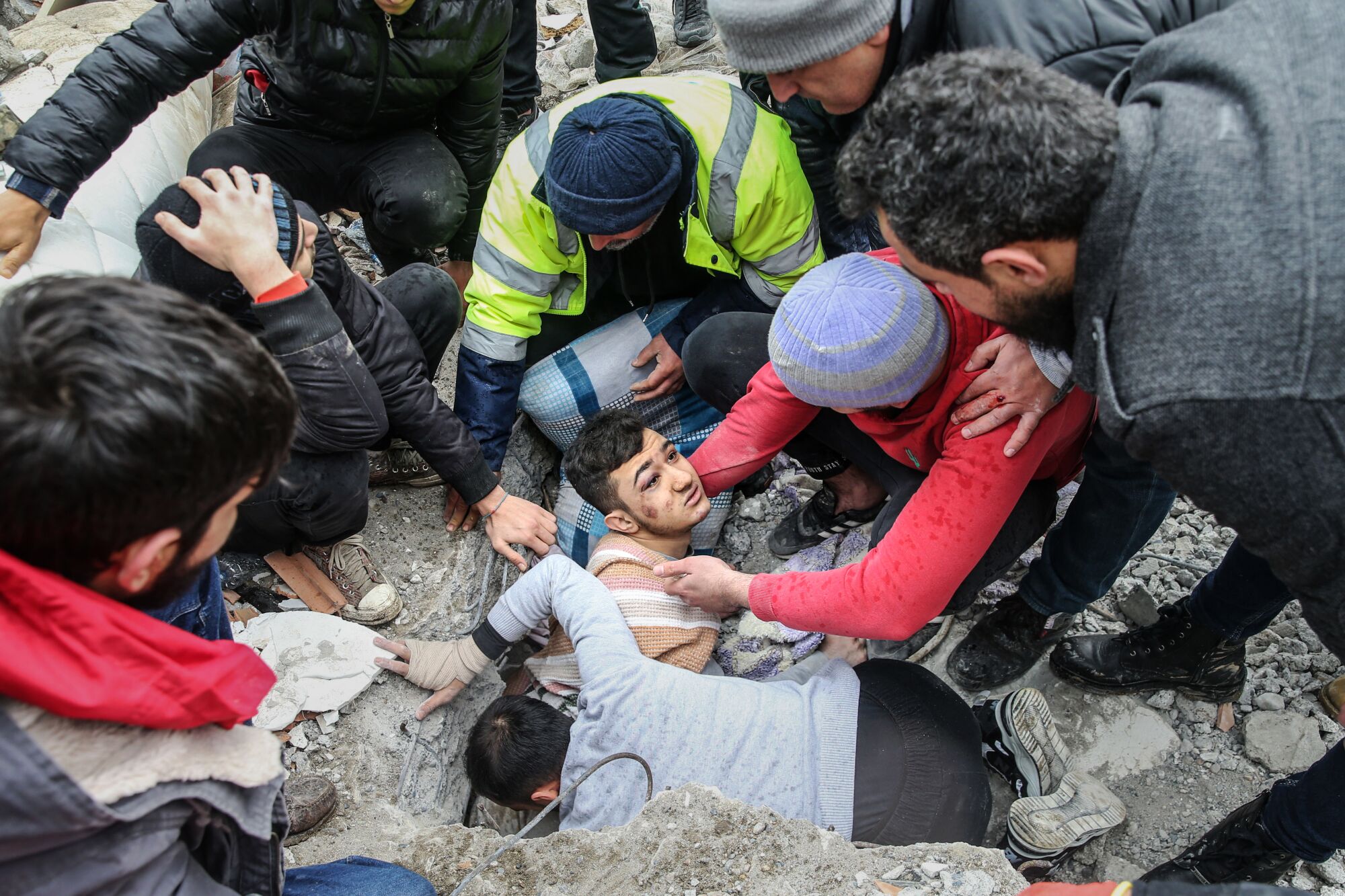 In a top-down-view a young man emerges, head and shoulders, from under rubble, being pulled out by a group of men.