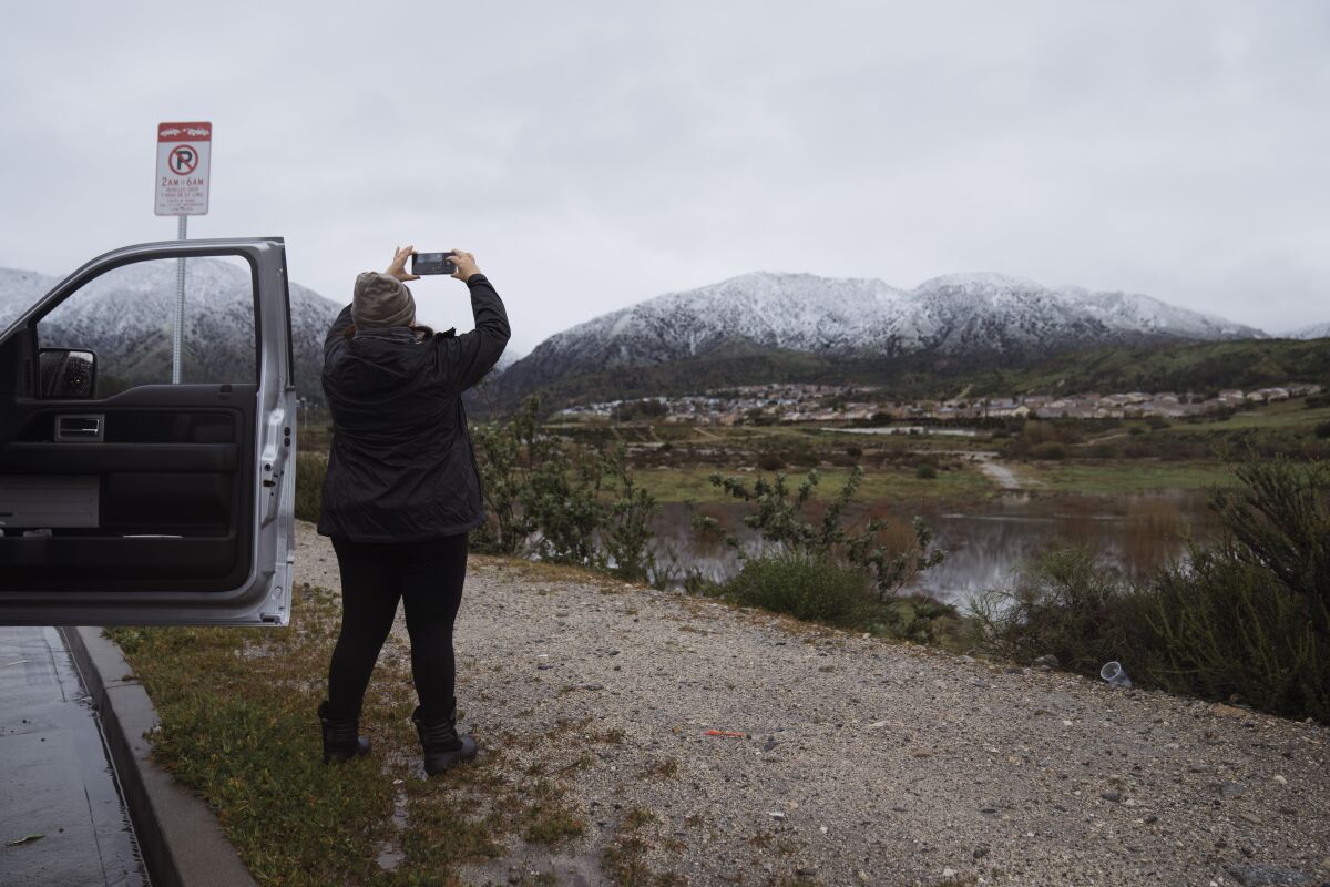 A woman stands beside a vehicle on the side of a road and photographs a snow-capped mountain on the horizon.