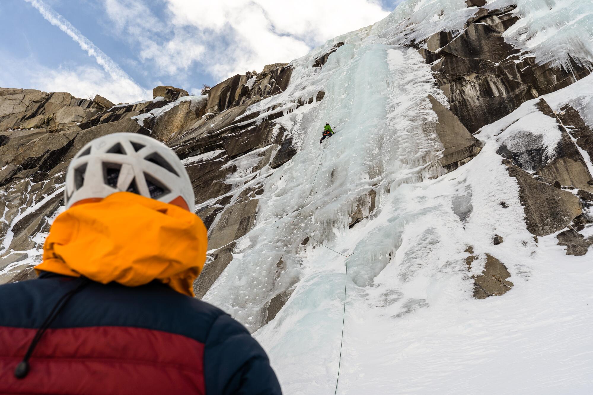 Seen from behind, a man wearing a helmet looks up at a frozen waterfall hugging a cliff. 