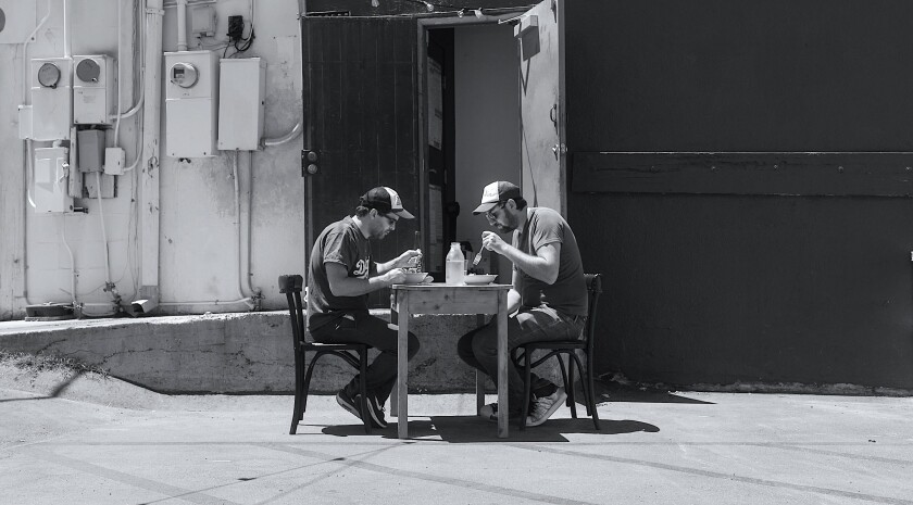 A black-and-white photo of two men eating outdoors at a small table in an alleyway.