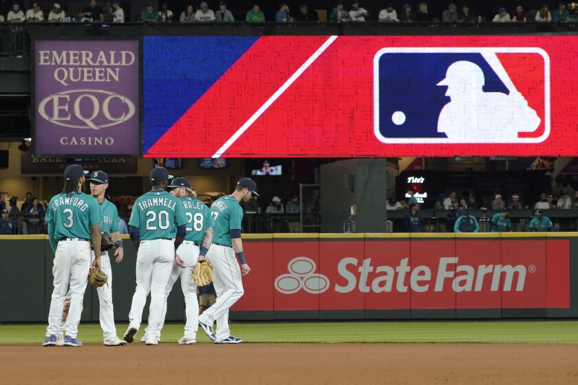 FILE - Seattle Mariners gather as the MLB logo is shown during a review of an attempted catch by right fielder Mitch Haniger of a ball hit by Tampa Bay Rays' Ji-Man Choi that was originally called an out during the ninth inning of a baseball game Friday, June 18, 2021, in Seattle. The call was overturned. While Major League Baseball and the players union try to chart a path forward, hoping to get baseball back on the field, some experts are watching the talks more from an academic viewpoint. (AP Photo/Ted S. Warren)