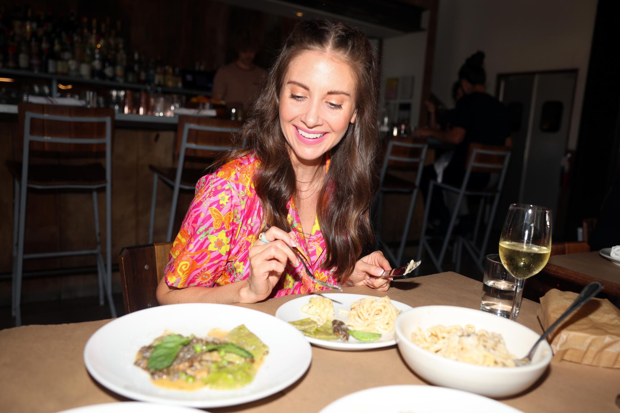 A woman with long hair sits at a table in a restaurant with three bowls of pasta in front of her.