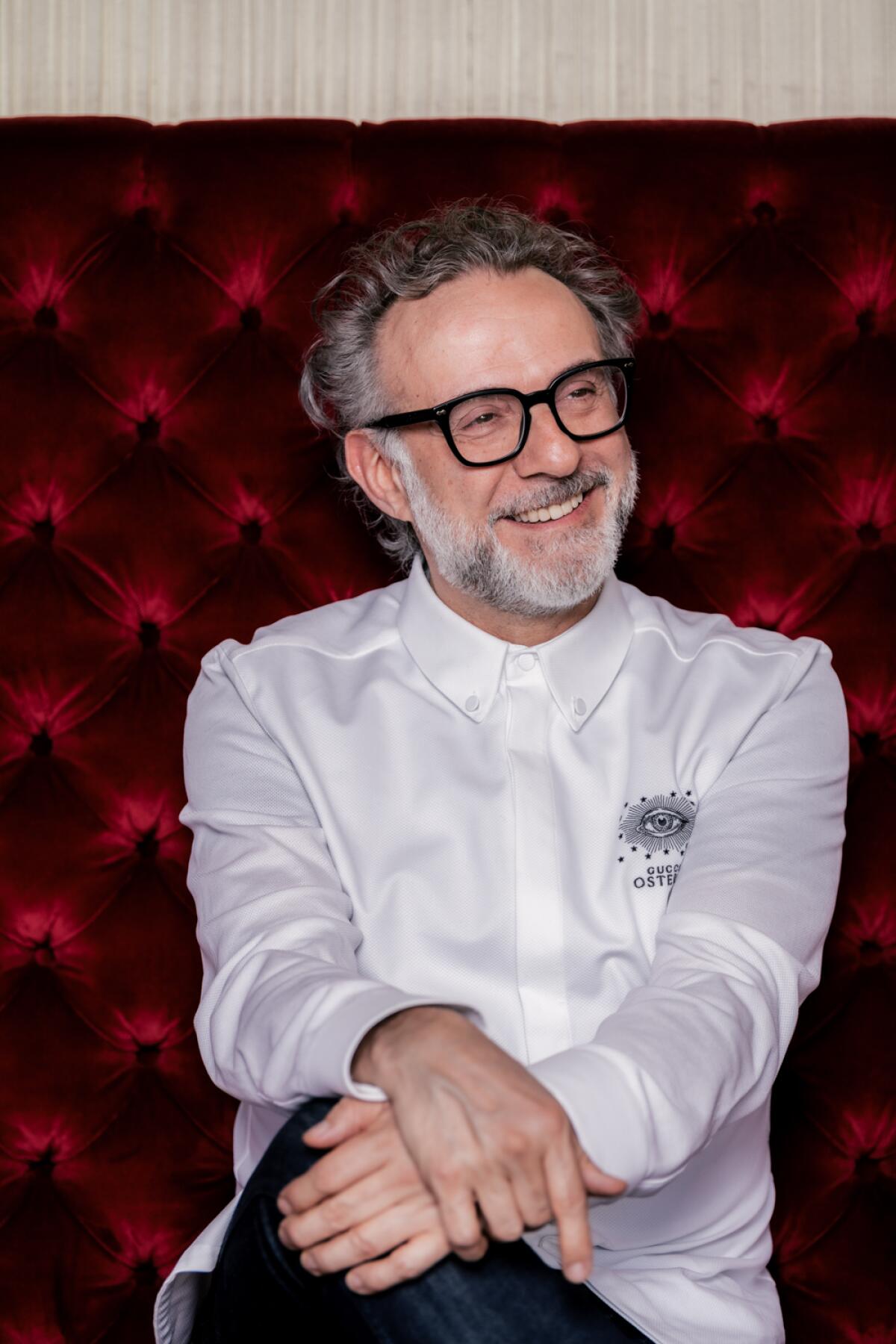 A chef, wearing dark-framed glasses, sits smiling with his hands crossed on his knee