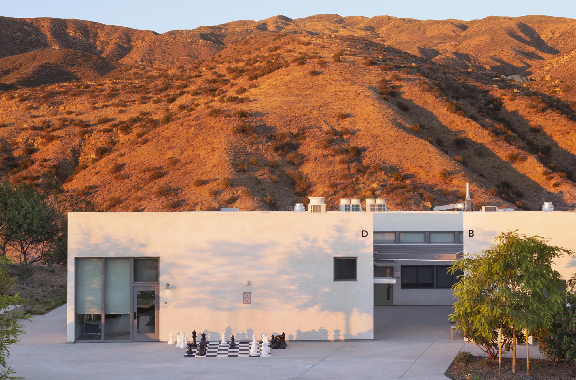 A minimalist white building with a giant chessboard in front sits before a large hilltop covered in Mediterranean scrub.
