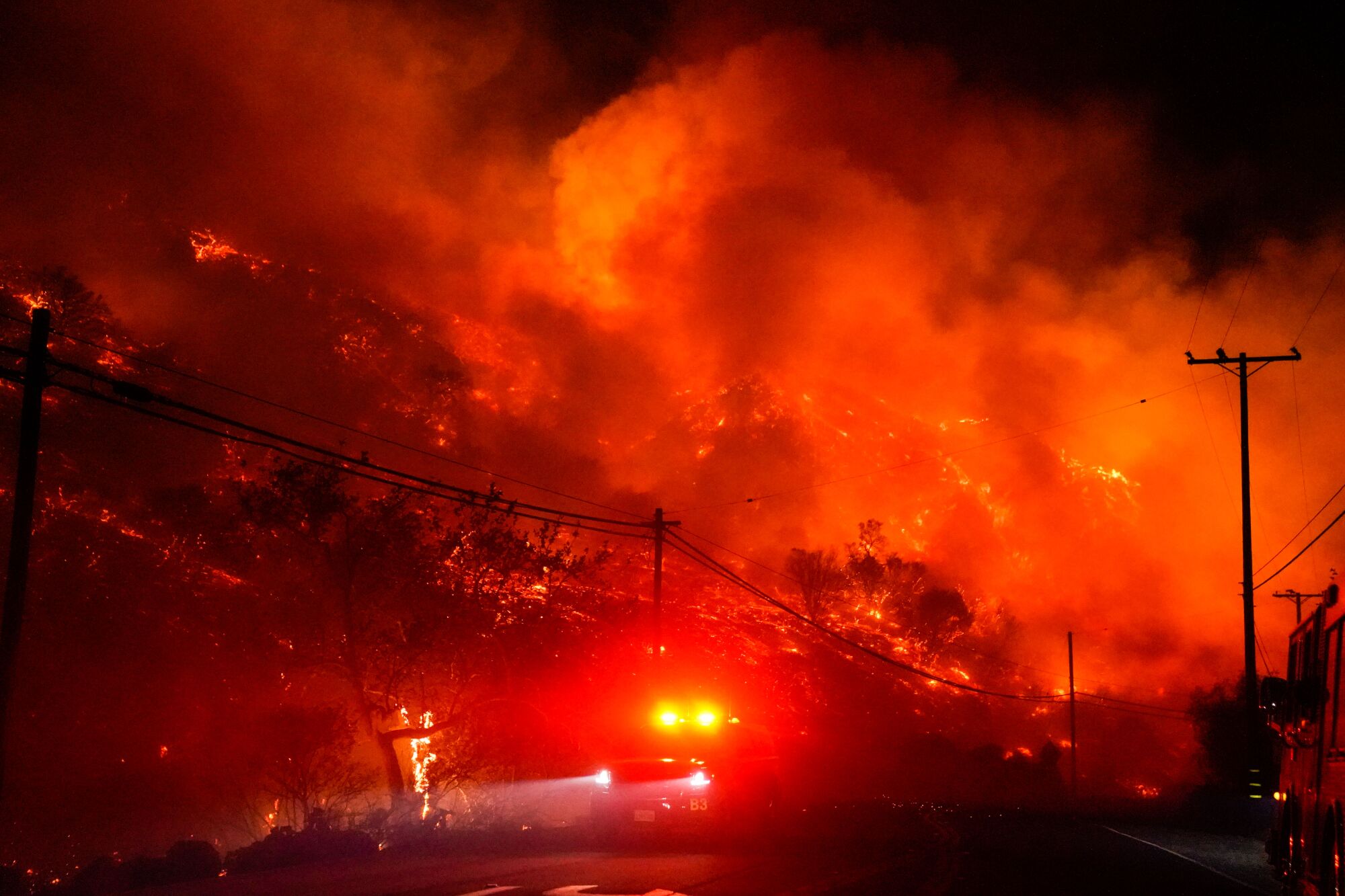 A fire truck's lights shine as it drives on a road at night with orange flames and embers on a hillside