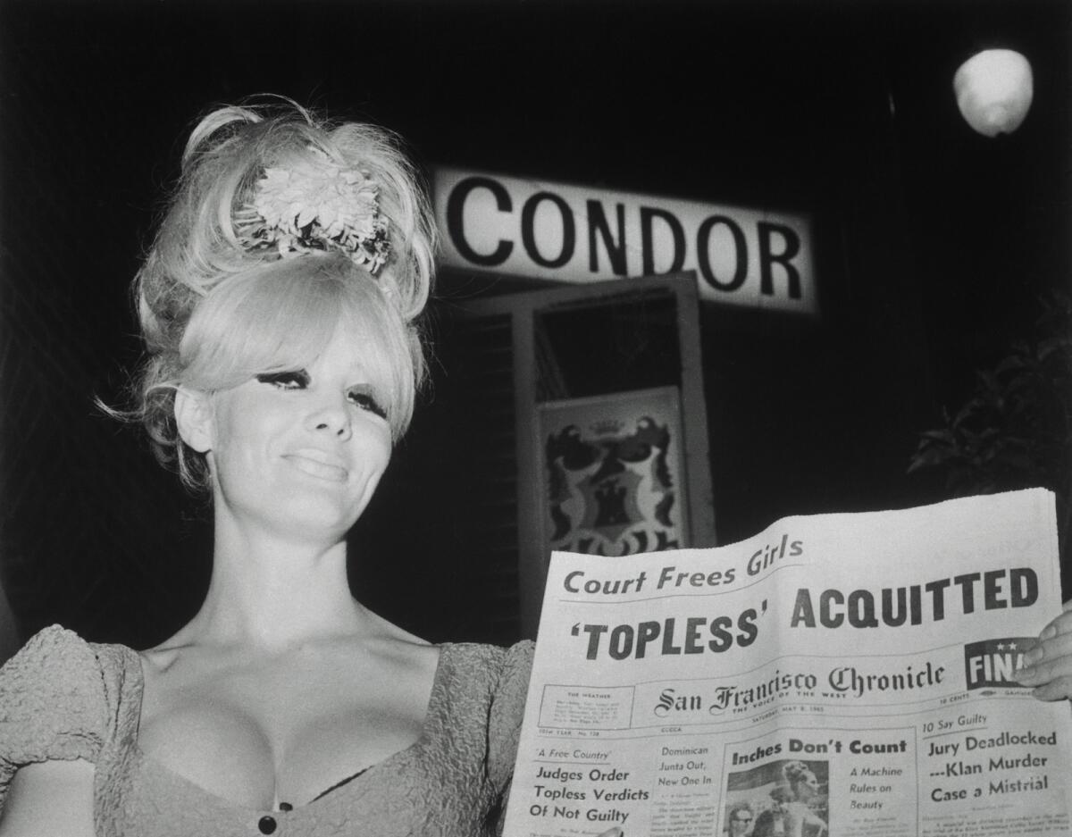 A woman holds a newspaper with the headline 'Topless' acquitted. A lighted sign that says Condor is behind her