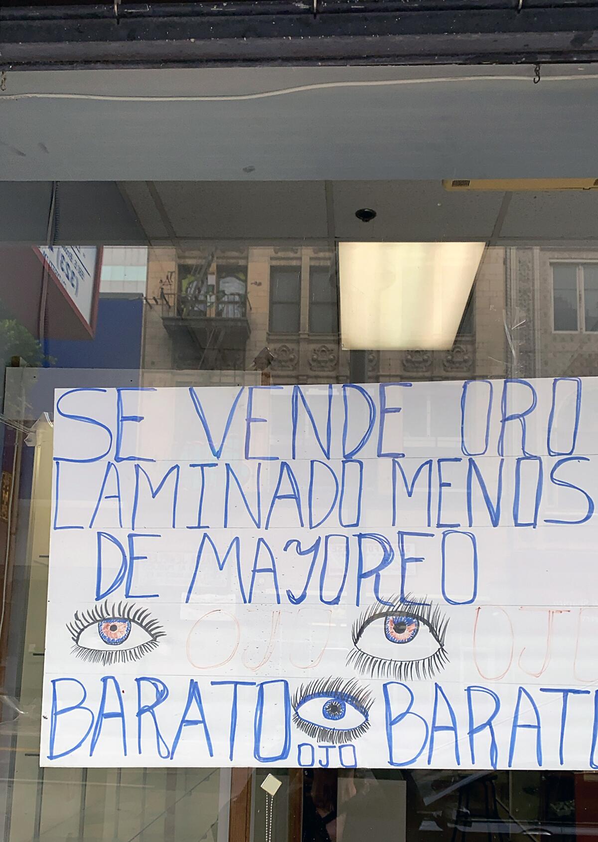 A handmade sign with eyes drawn throughout reads "Se Vende Oro Laminado Menos de Mayoreo — Barato Ojo Barato"
