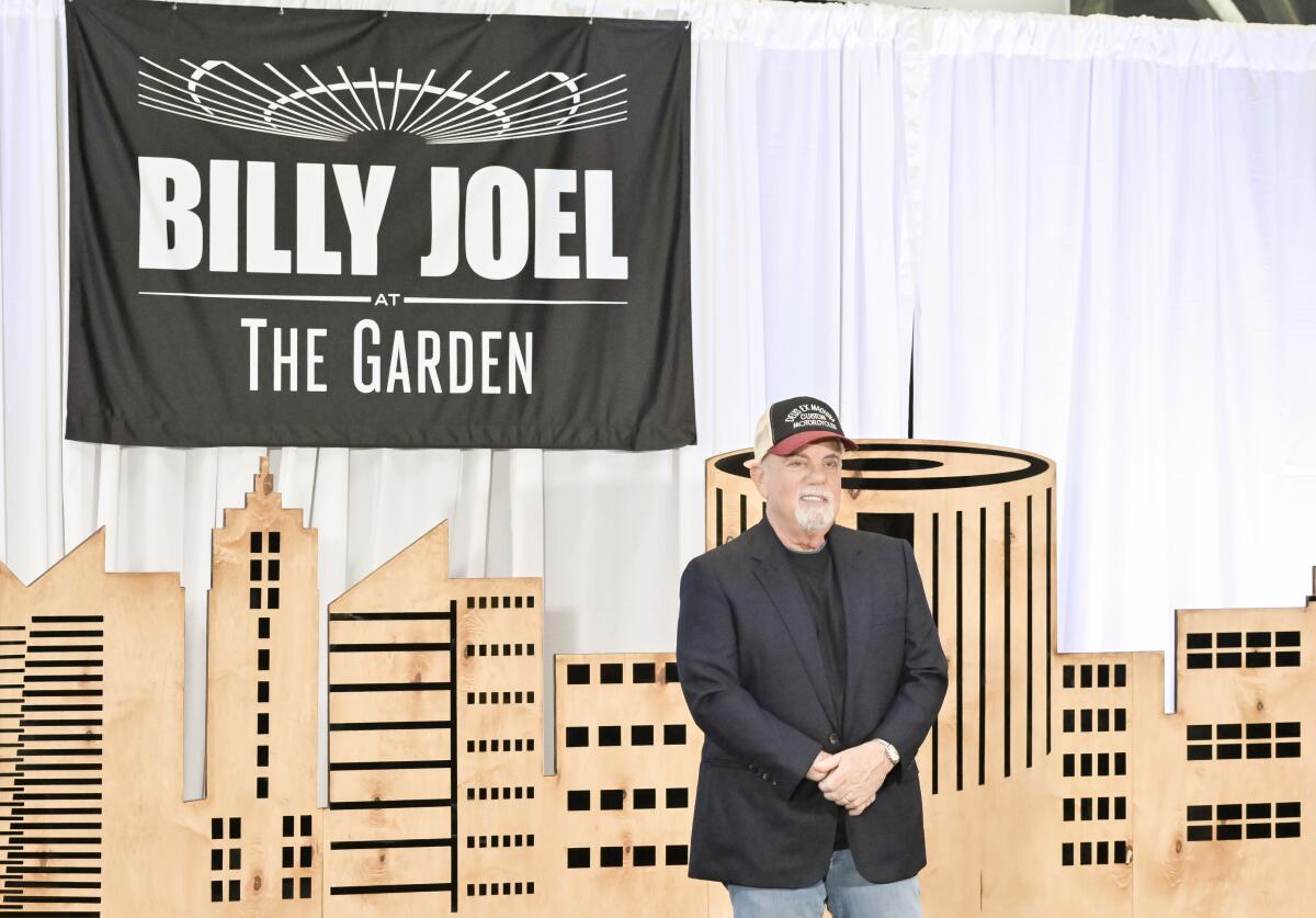 Billy Joel, in black blazer, jeans and ball cap, poses in front of a cloth backdrop that says "Billy Joel: The Garden."