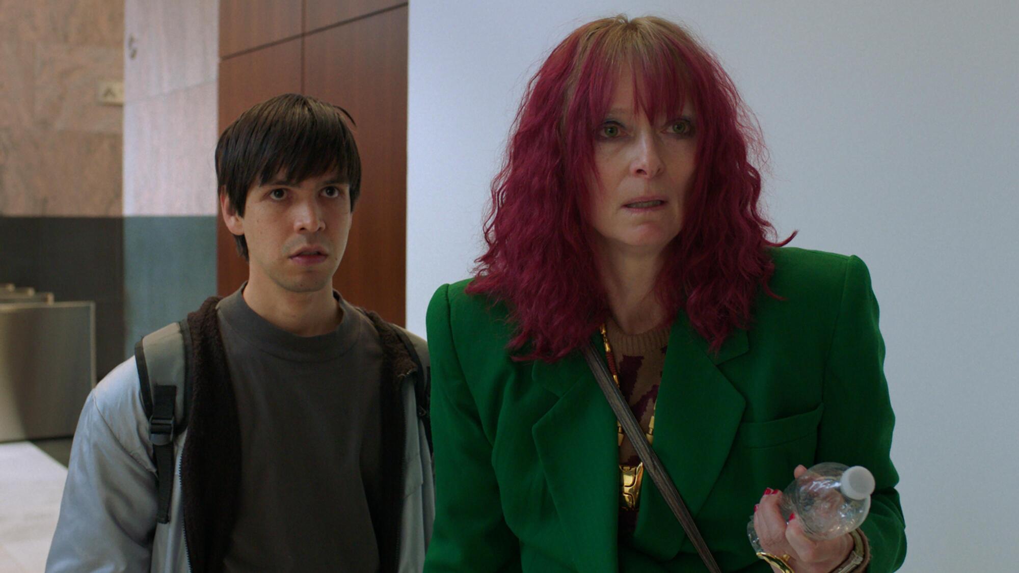 An assistant in a dowdy sweatshirt and backpack accompanies his red-haired boss.