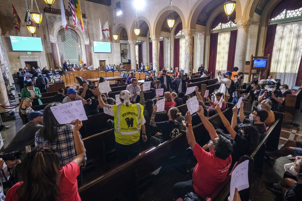 Observers hold signs in protest in L.A.'s architecturally ornate City Council Chamber.