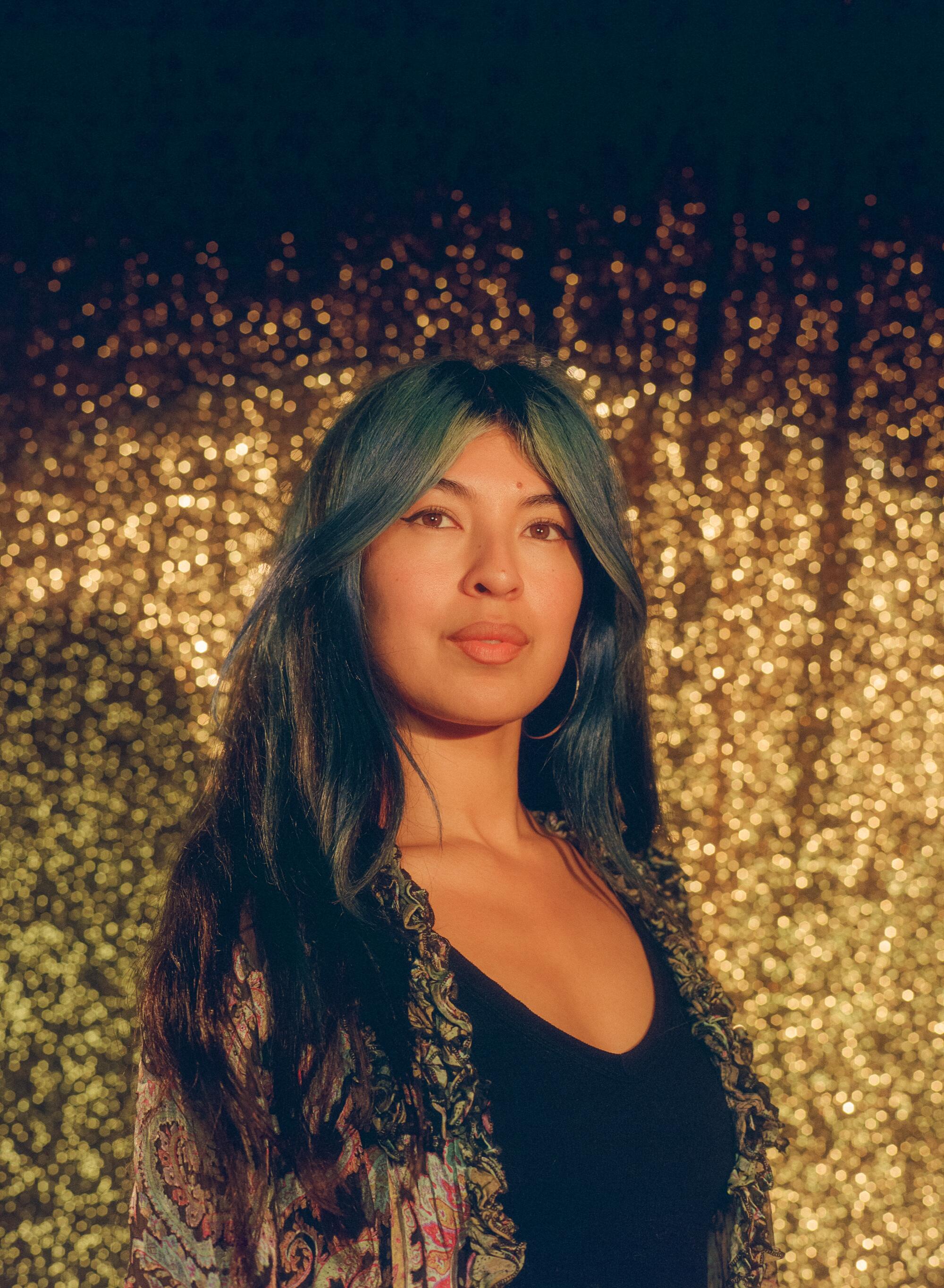 A woman in blue hair stands in front of a golden curtain.