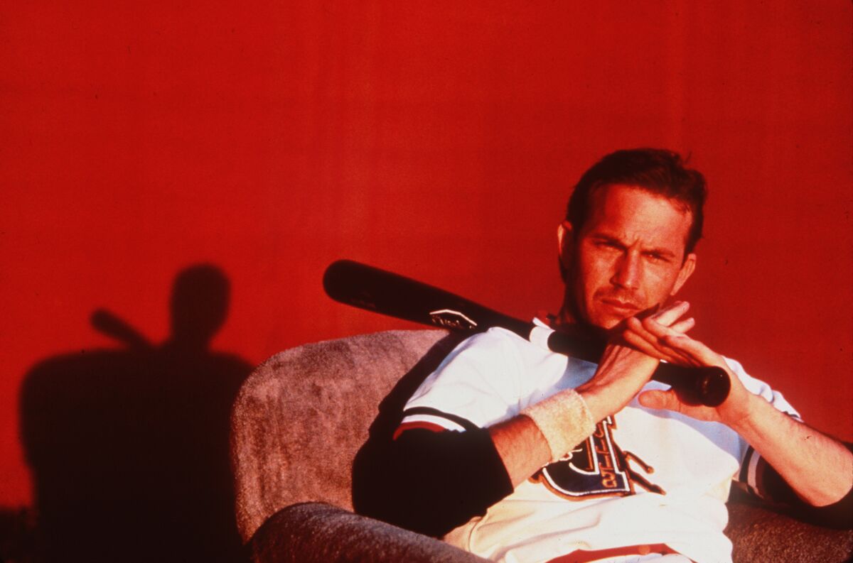 A man in a baseball uniform sits in an armchair, resting a baseball bat on his shoulder.