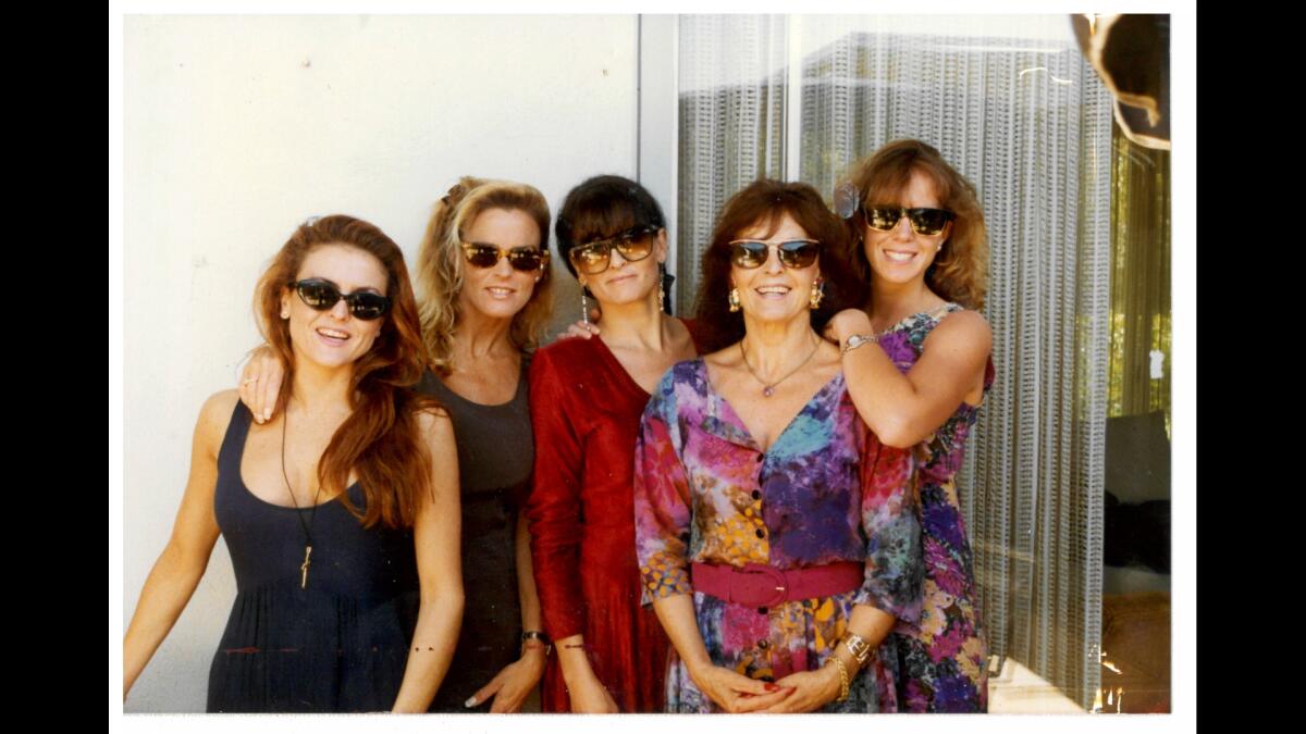 Five women in sunglasses standing by a doorway with their hands on each other's shoulders.