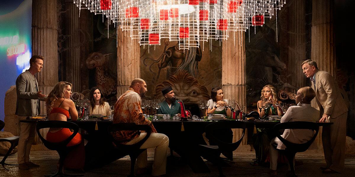A group of people sit around a large table in a scene from "Glass Onion: A Knives Out Mystery." 