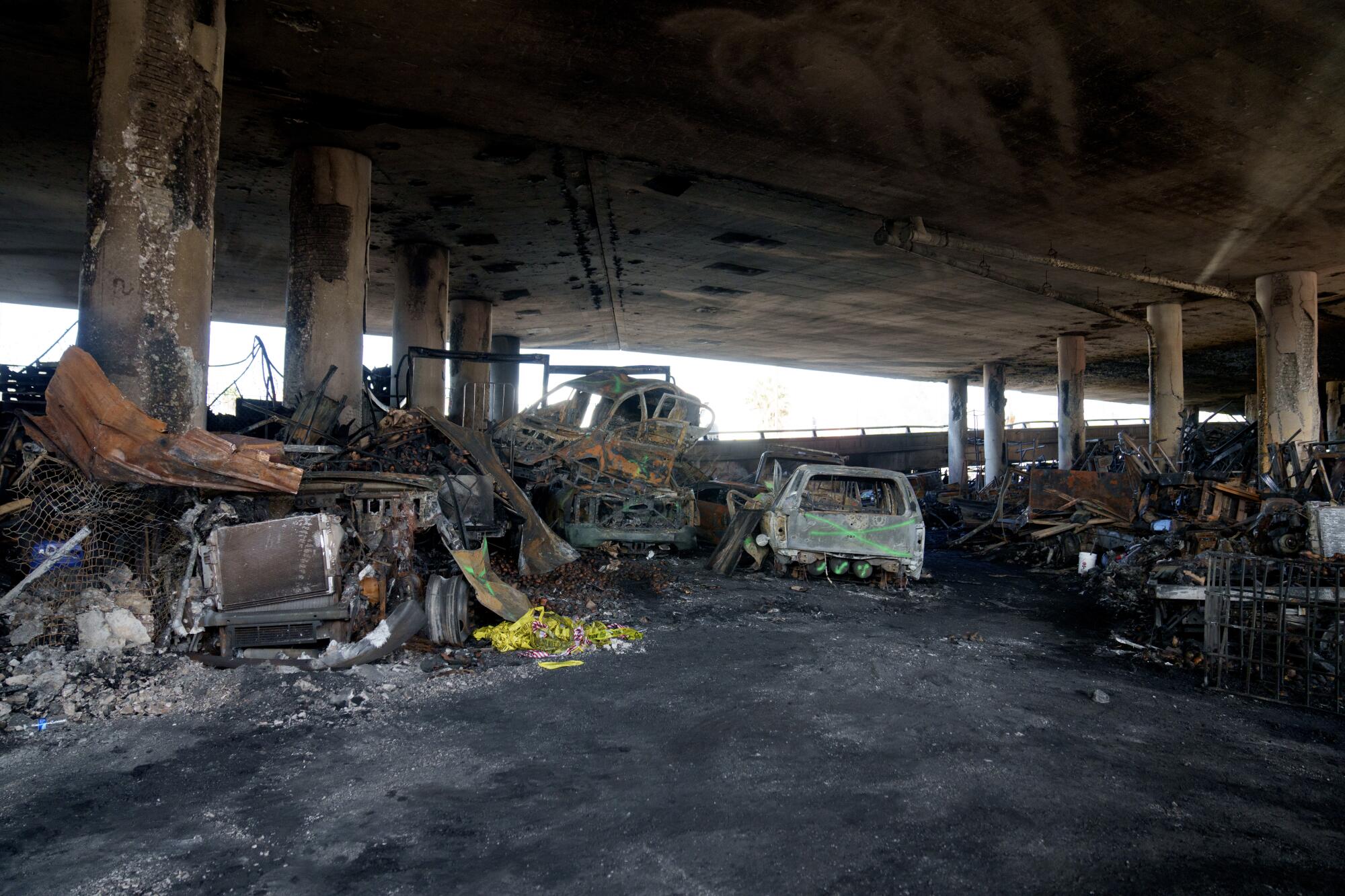 Burned debris including vehicles piled next to the charred support posts underneath a freeway.
