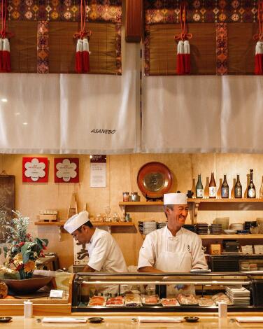 Three people at a sushi bar, with three smiling chefs standing behind the bar