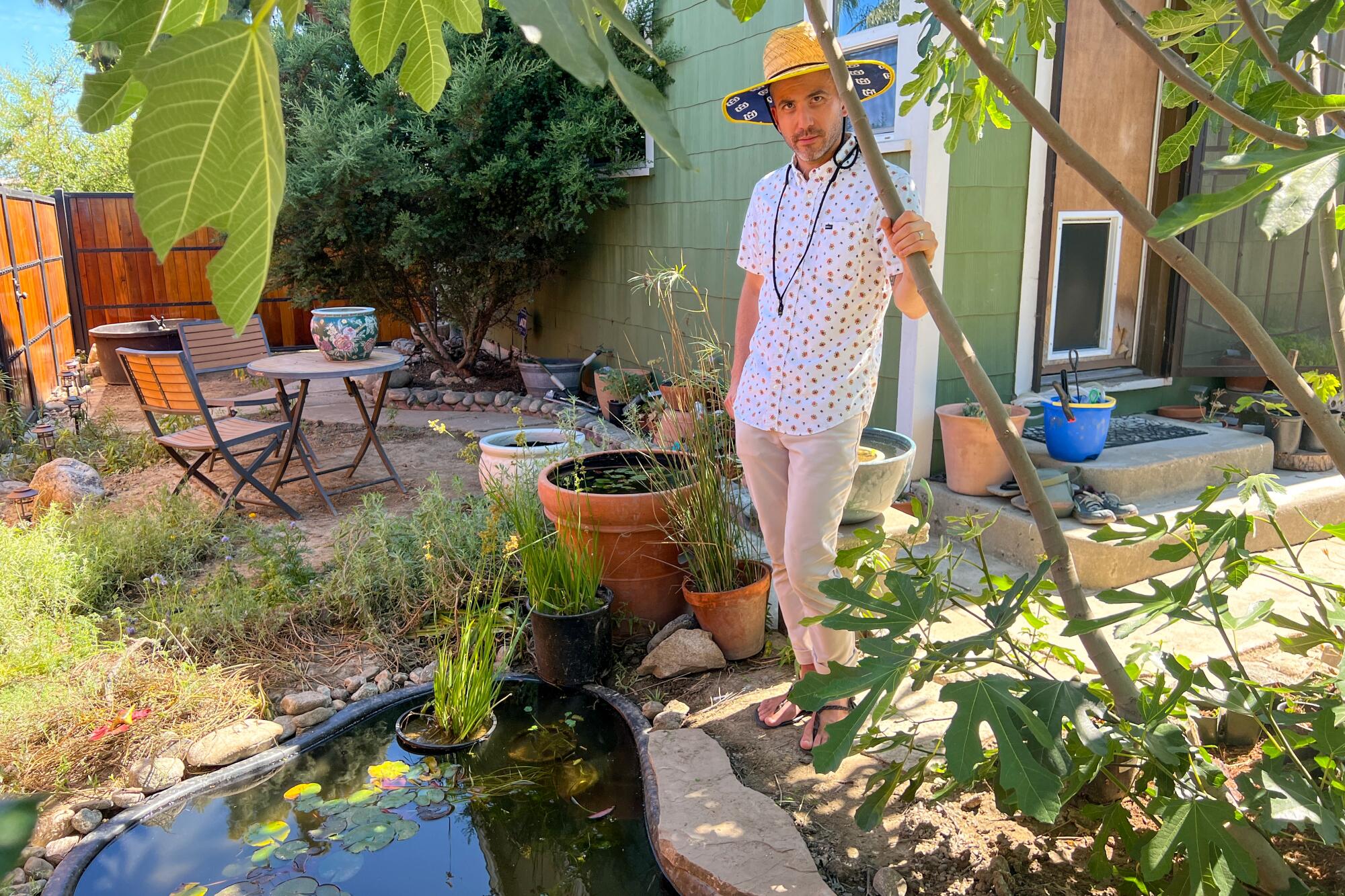 Andrew Chaves, a slender man in a sun hat and sandals, stands next to the small pond he installed.