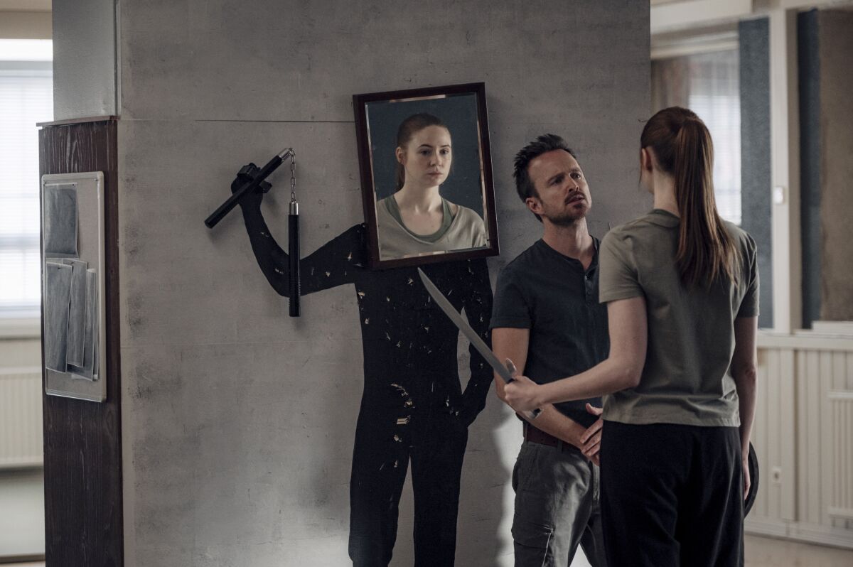 A woman holding a long knife is reflected in a mirror of the shoulder of a man facing her.