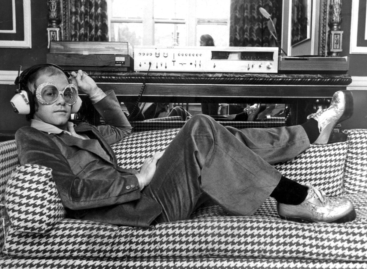 A young Elton John reclines on a couch listening to music on headphones