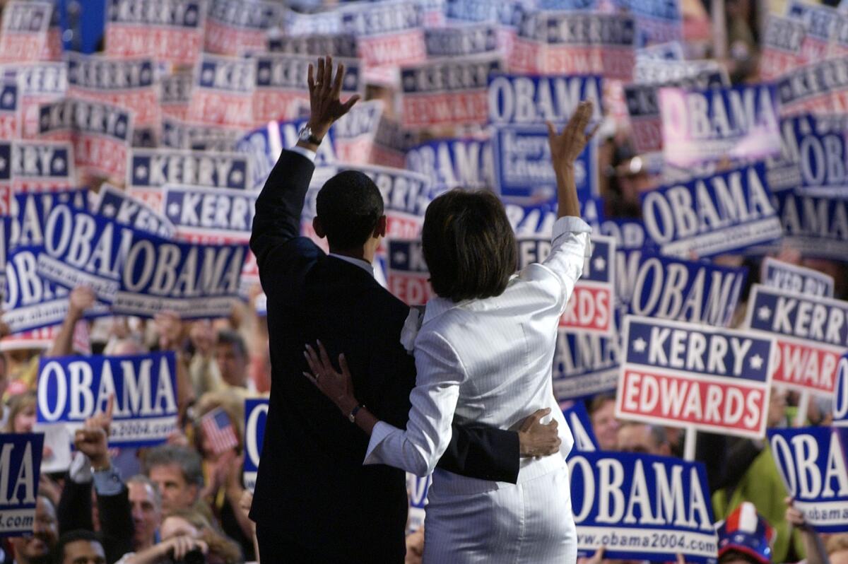 Barack and Michelle Obama seen from behind, an arm around each other's waist, waving to a crowd of people holding signs.