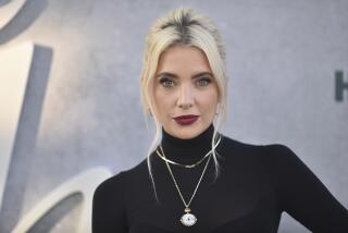 Ashley Benson, with blond hair and sporting a dark red lip, poses in a long-sleeve black turtle neck and thin gold necklaces.