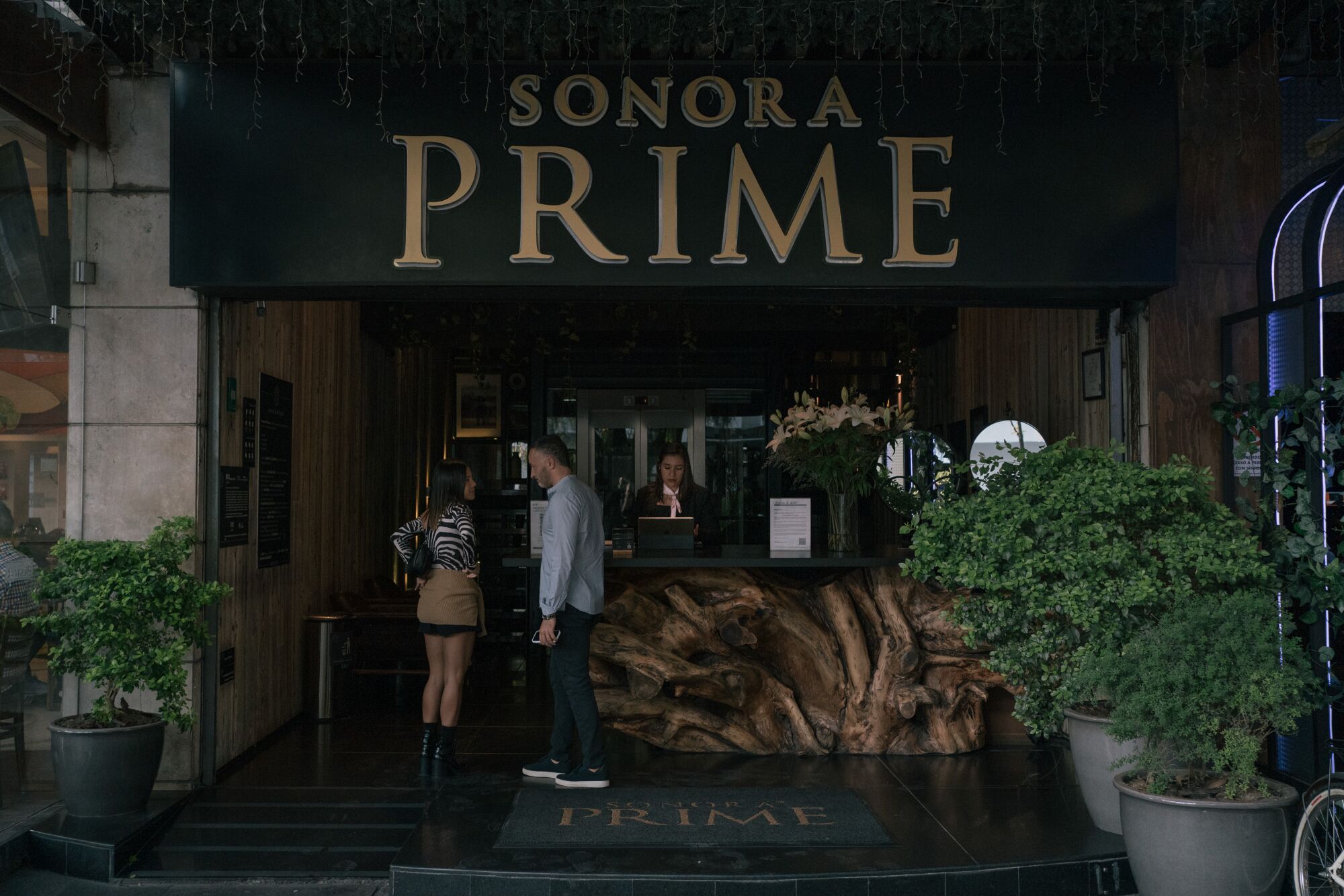 A woman, left, and a man stand in front of a reception area flanked by plants and a sign overhead that says Sonora Prime
