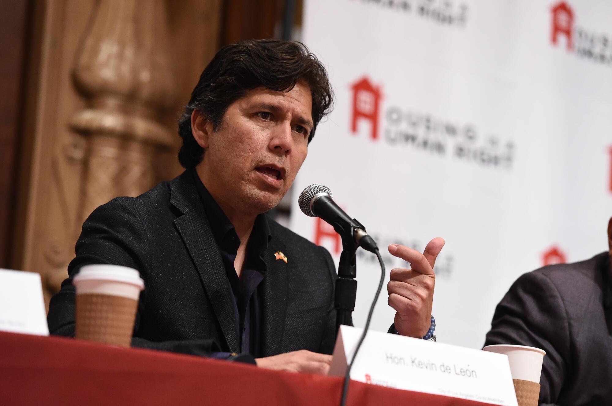 Kevin de Leon, speaking into a microphone, seated behind a red-covered table.