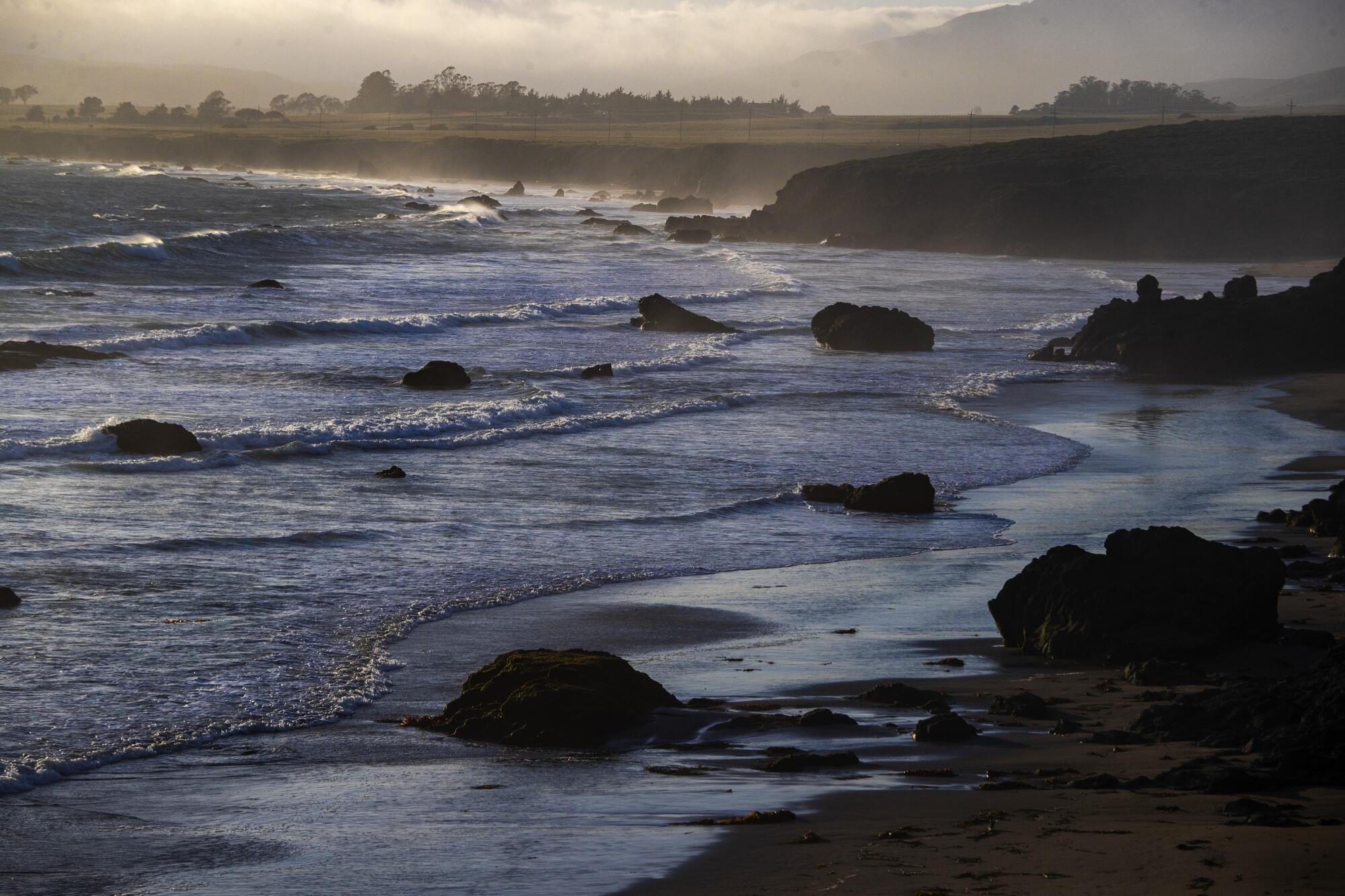 A view of flat, deserted beach and ocean surf rolling in over low boulders.