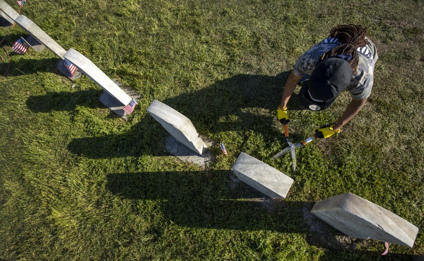 A woman with shears clips grass around a gravestone.