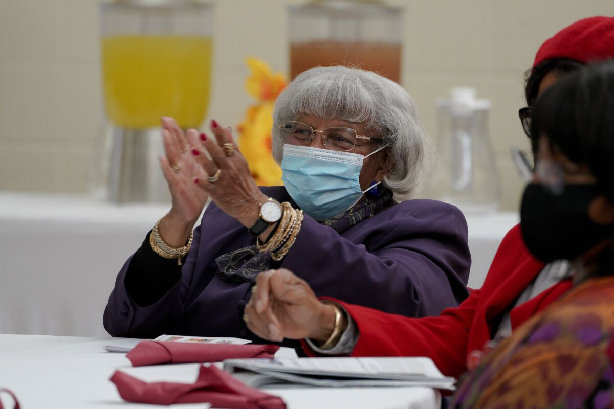 a woman with gray hair in a purple blazer wearing a blue medical mask, watch, glasses and gold bracelets and rings claps.