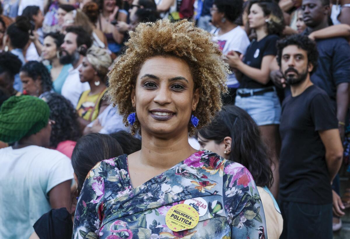 A woman with curly, short brown hair, in a blue-and-pink patterned top, smiles while standing in a crowd