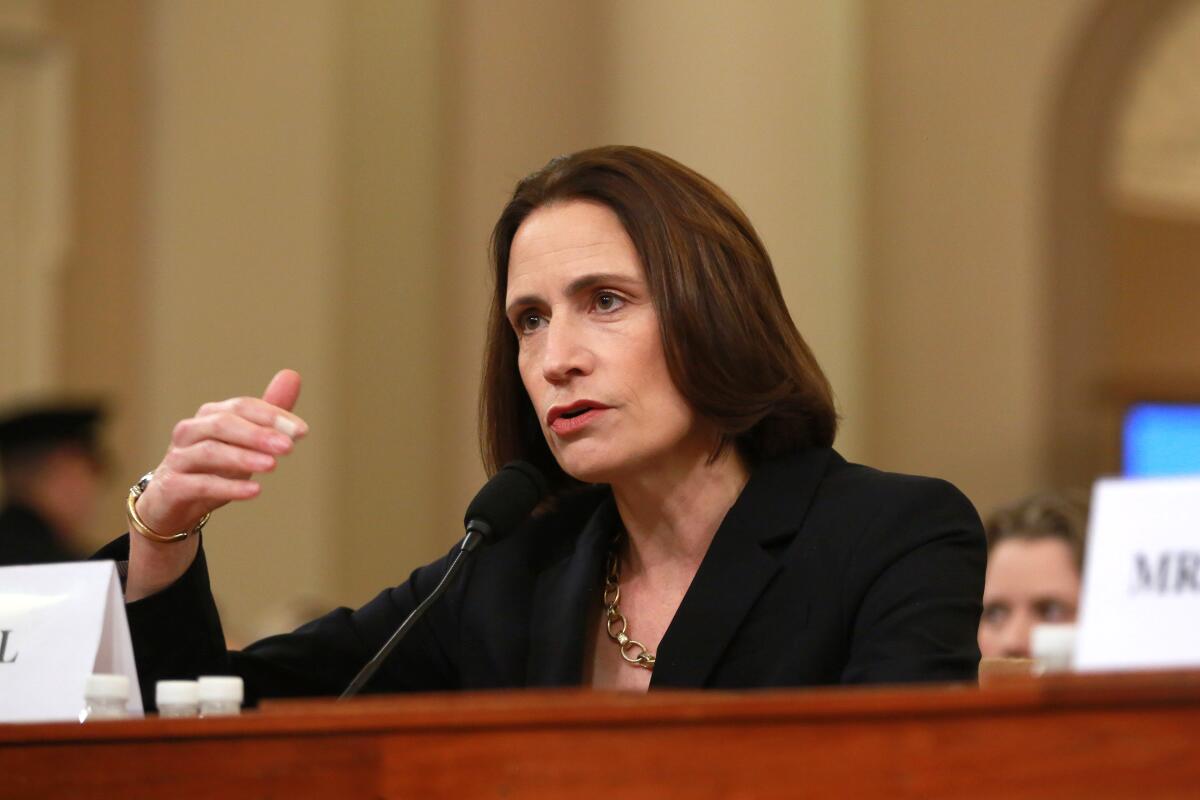 A woman with dark hair, in a dark suit, gestures with her hand while speaking at a mic