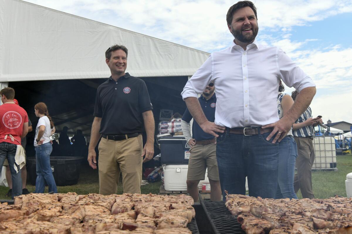 A bearded man, in white shirtsleeves, stands with hands on hips near another man, also smiling