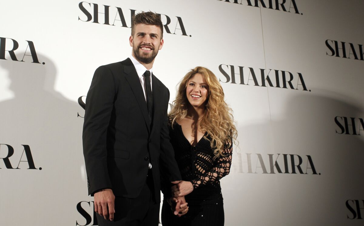 A tall man stands next to a short blond woman while standing in front of a Shakira backdrop. 