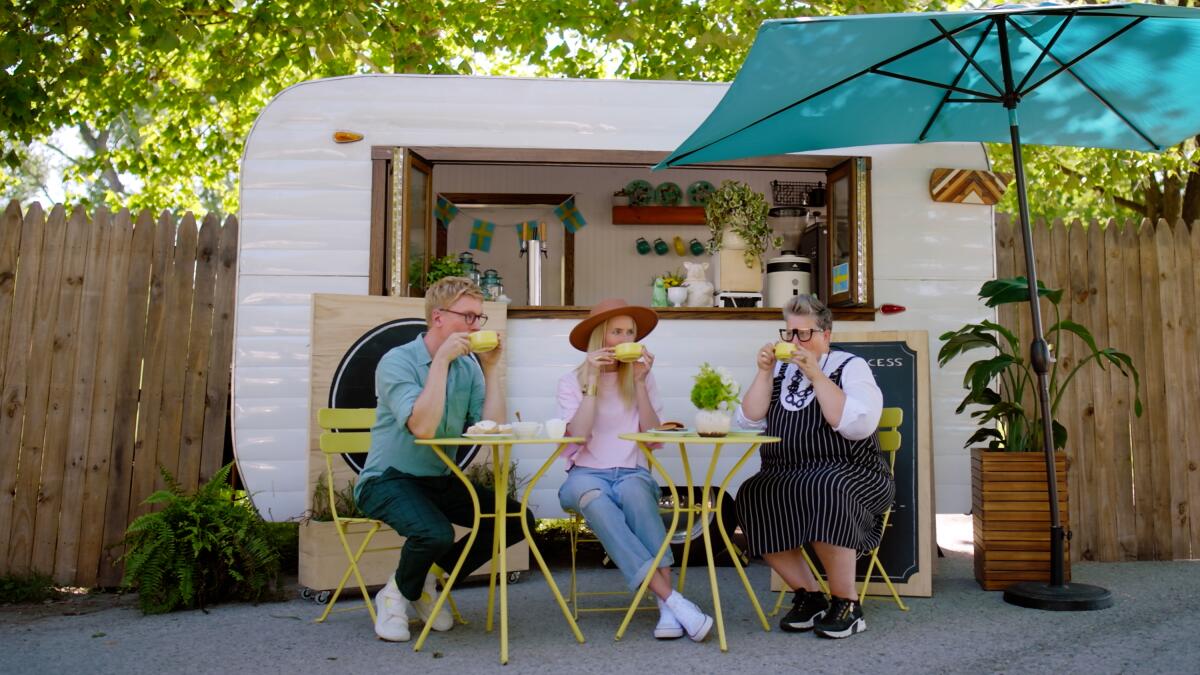 Three Swedish people sit at a bistro table drinking coffee outside a coffee cart.