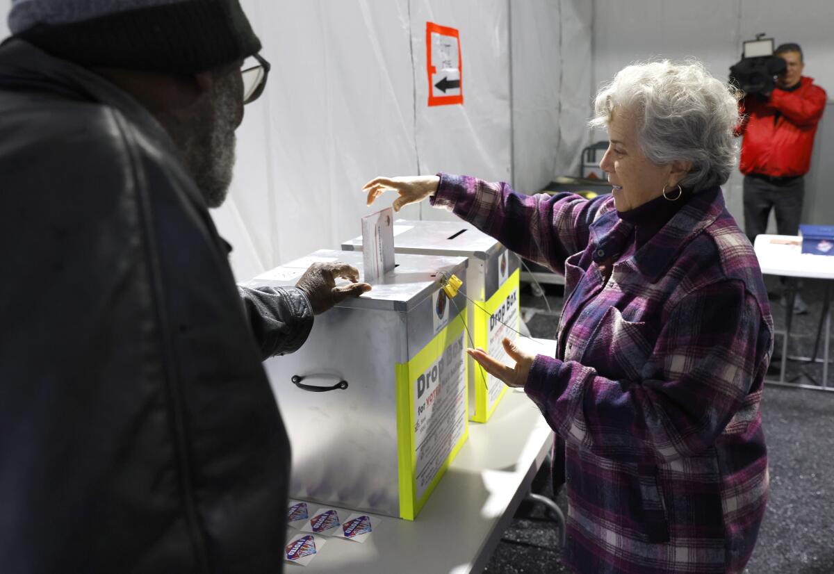 A woman in a plaid purple jacket puts a ballot into a silver box.