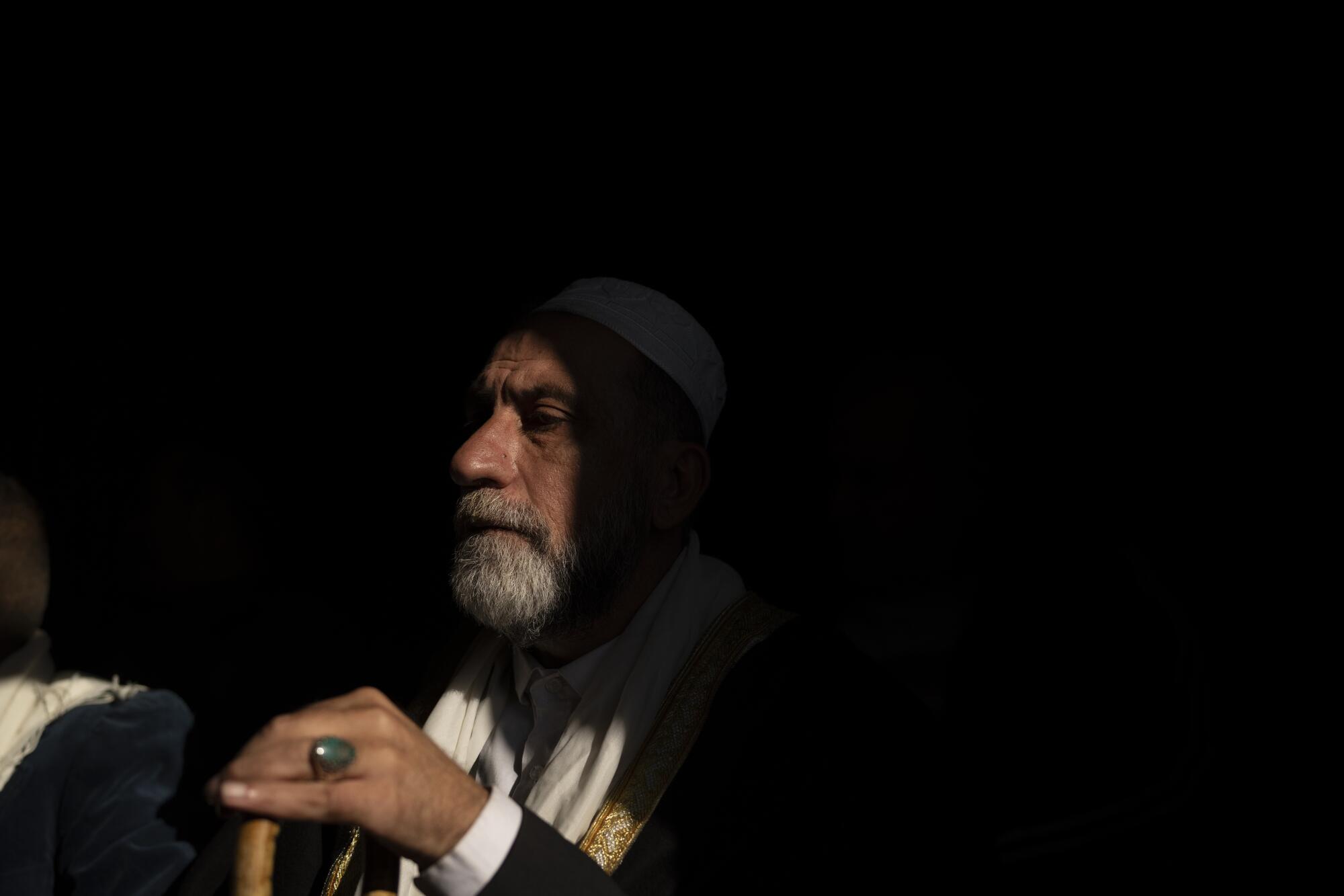 A man with a gray beard and white skullcap is shown at a meeting