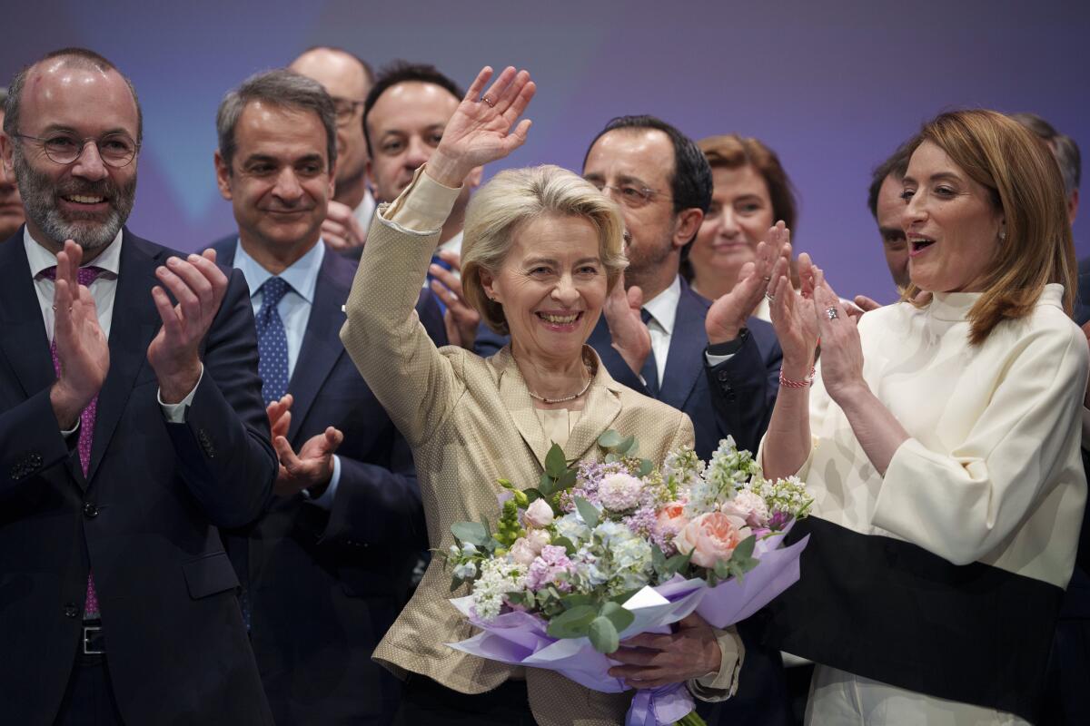 European Commission President Ursula von der Leyen holds flowers and waves as people around her applaud.