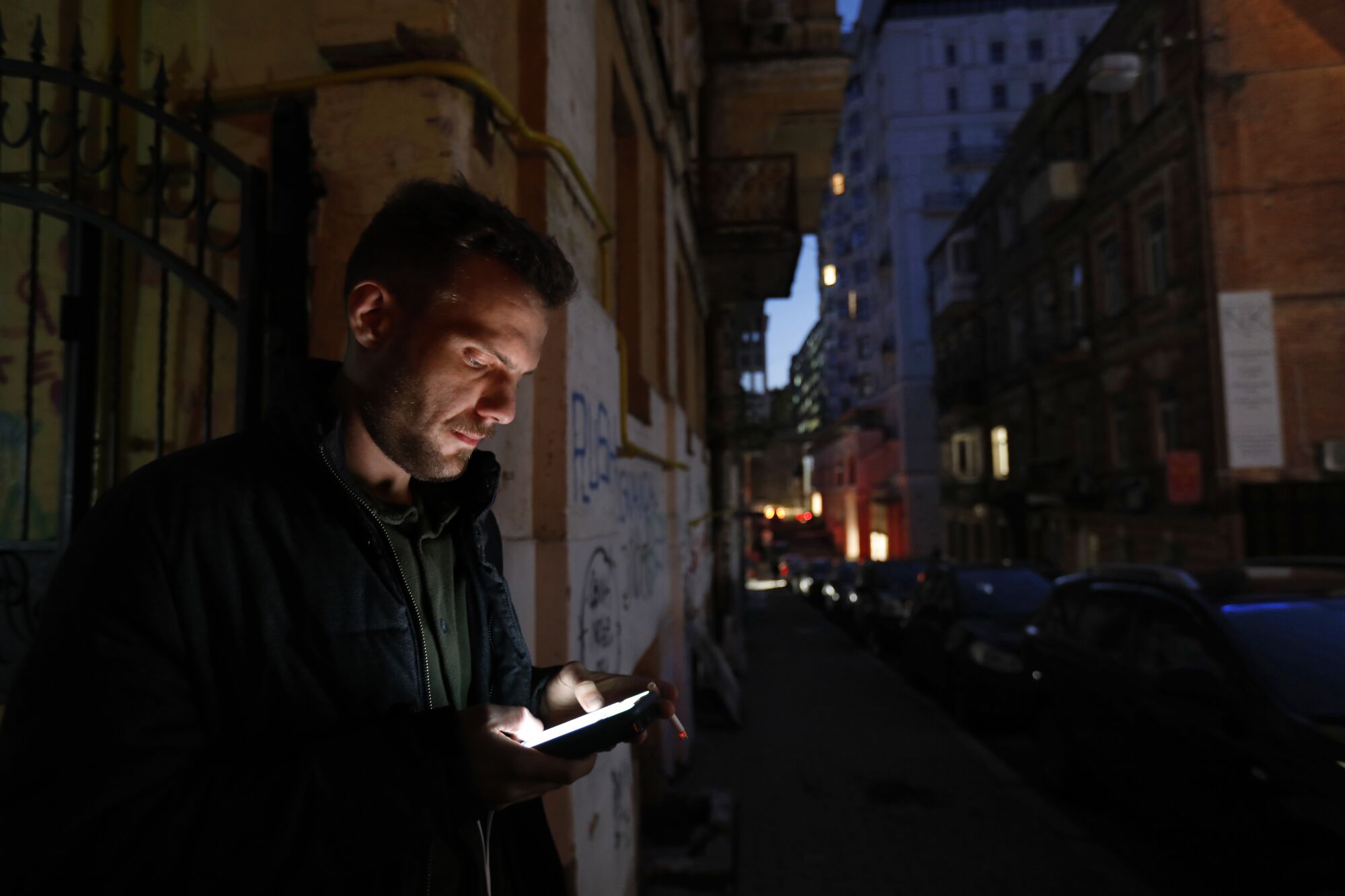 A man's phone lights up his face as he stands on a darkening city street