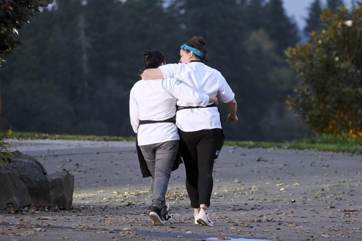 Two women in chef's coats walk away from the camera, arms around each other.