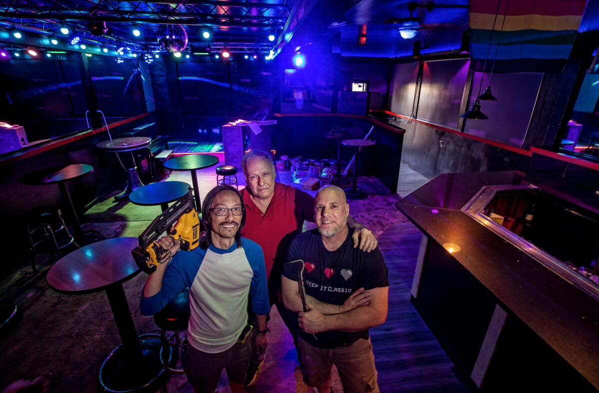 Three men stand inside a dark, blue-lit bar.