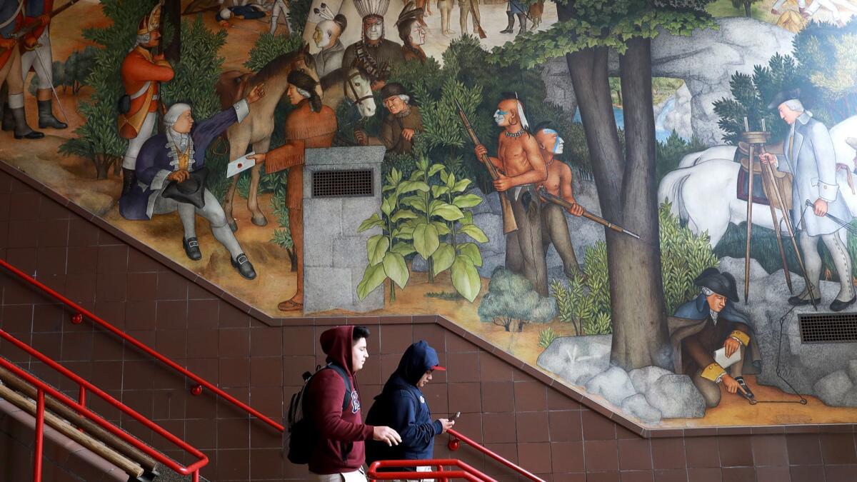 Students walk down stairs in front of a high school's mural of Native Americans and white settlers