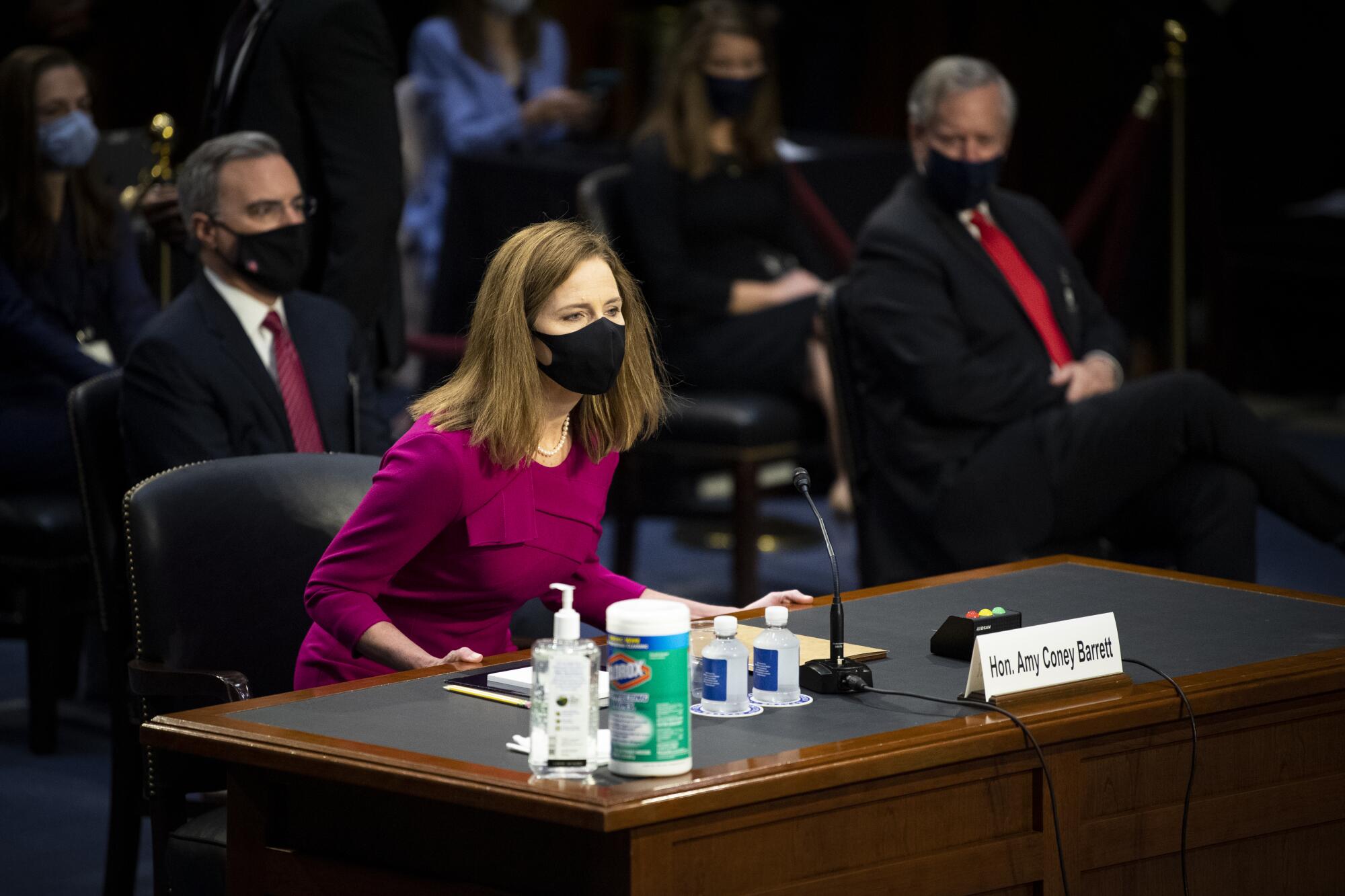 Amy Coney Barrett, masked, takes a seat behind a desk that has water, hand sanitizer and Clorox wipes.