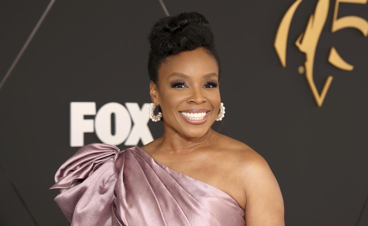 Amber Ruffin with her hair in an updo, wearing a mauve one-shoulder dress and pearl hoop earrings. 