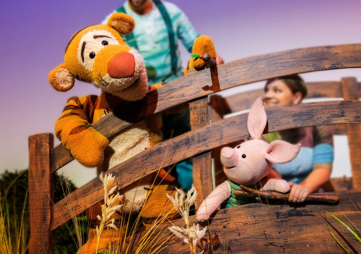 In a stage show, Tigger looks at Piglet, who peeks out between wood planks of a fence.