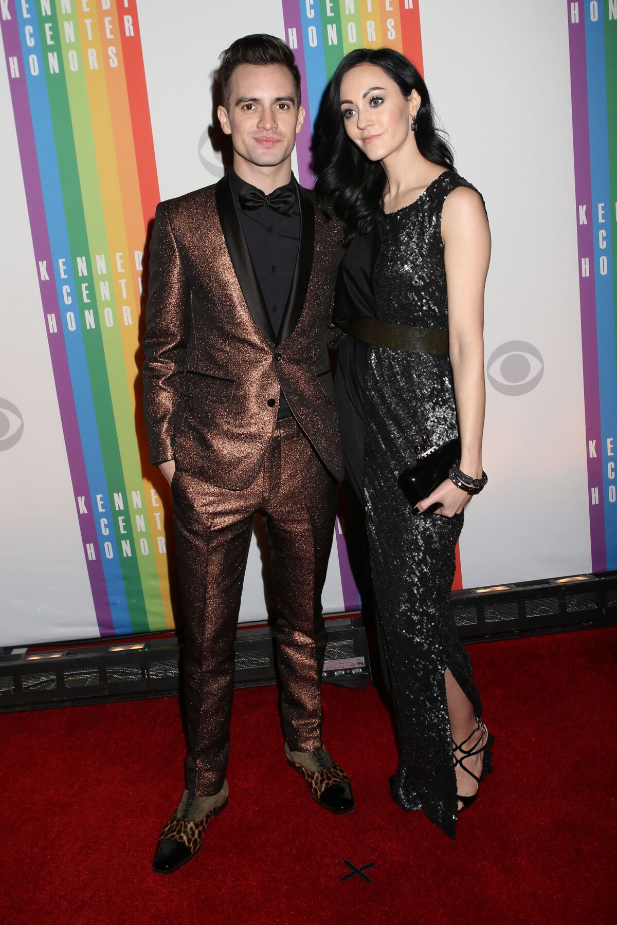 A man in a copper suit and a dark-haired woman in a dark gown pose on a red carpet