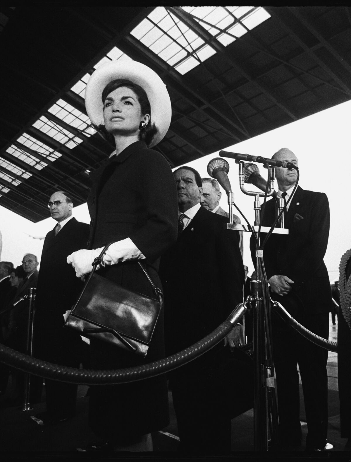 A woman in a white hat and gloves, wearing a dark suit and carrying a dark purse on her wrist.