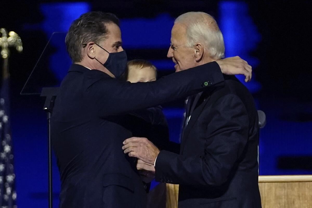 Two men in dark suits, one younger and wearing a mask on the left, embrace 